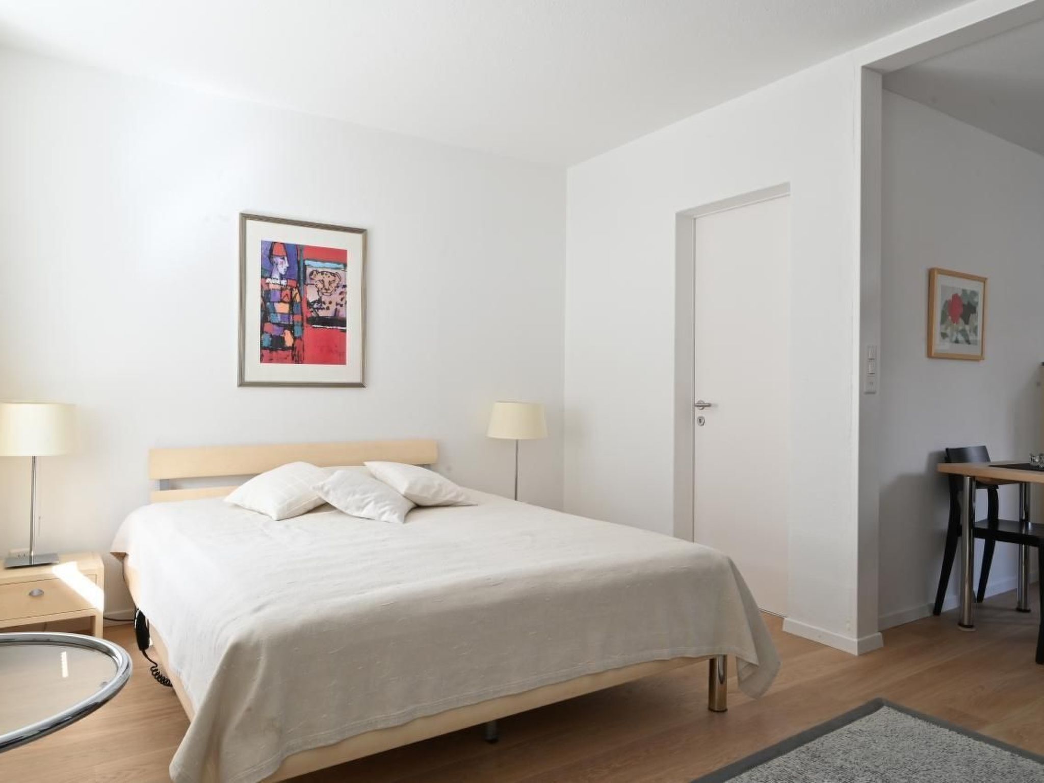 A bedroom with a bed, bedside lamps, a desk, and a door.