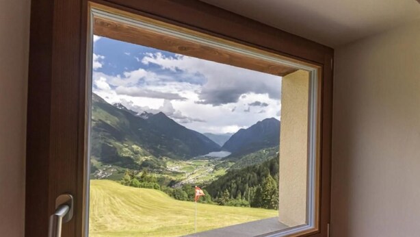 A room with a large window offering a view of a mountainous landscape with a lake and a flag. - Monte Campell Dasura by Interhome (Poschiavo)
