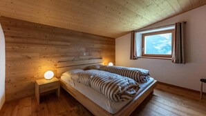 A cozy bedroom with a wooden ceiling, a bed with striped bedding, a nightstand with a lamp, and a window with curtains. - Monte Campell Dasura by Interhome (Poschiavo)