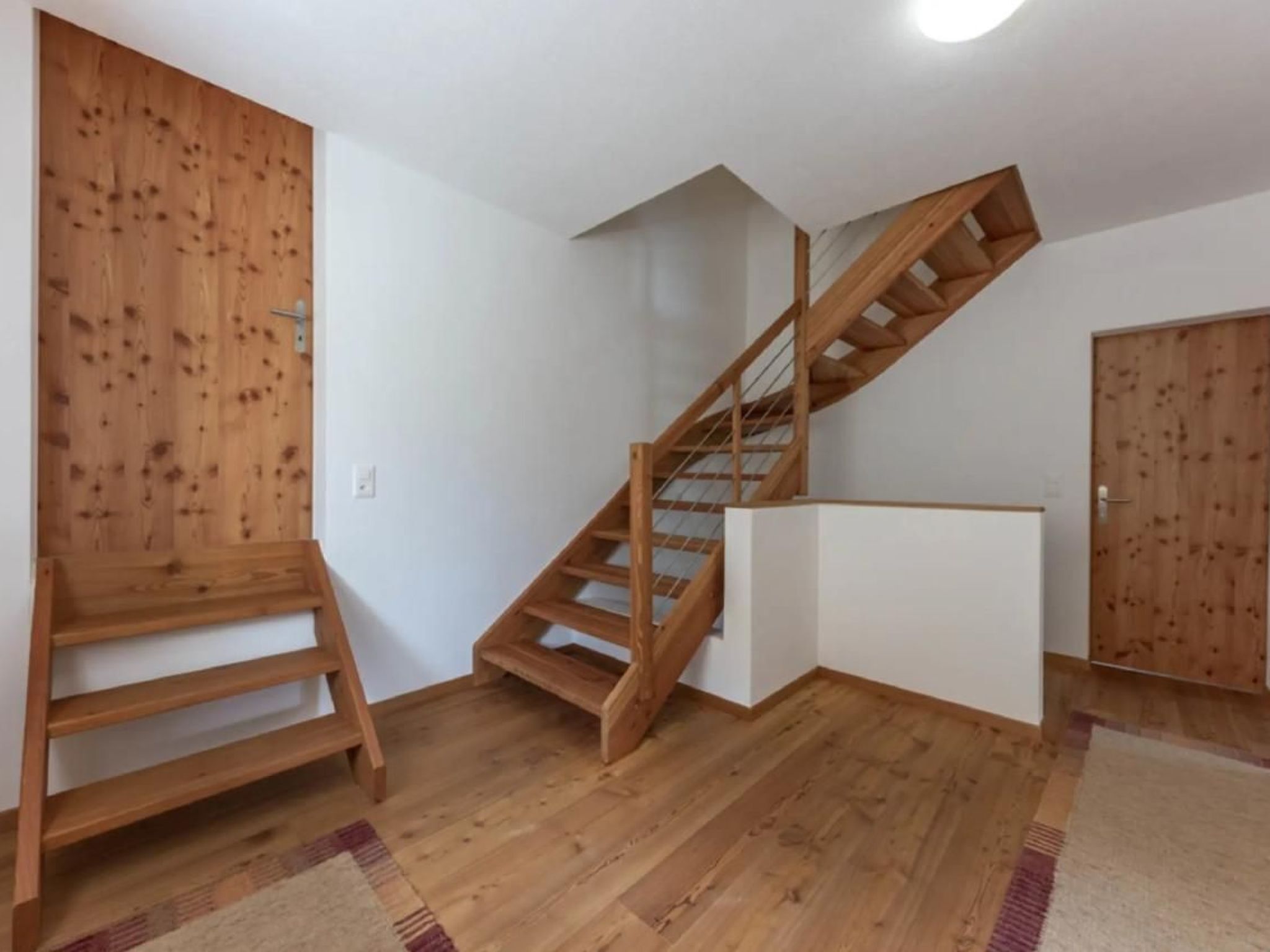 A staircase with wooden railings leading to a landing, a wooden bench, and a white wall with a door.