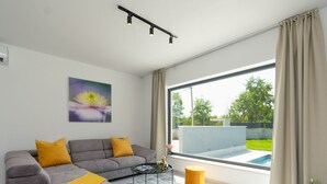 Una sala de estar moderna con un sofá gris, cojines amarillos, una mesa de centro y un amplio ventanal con vista a una piscina y follaje.