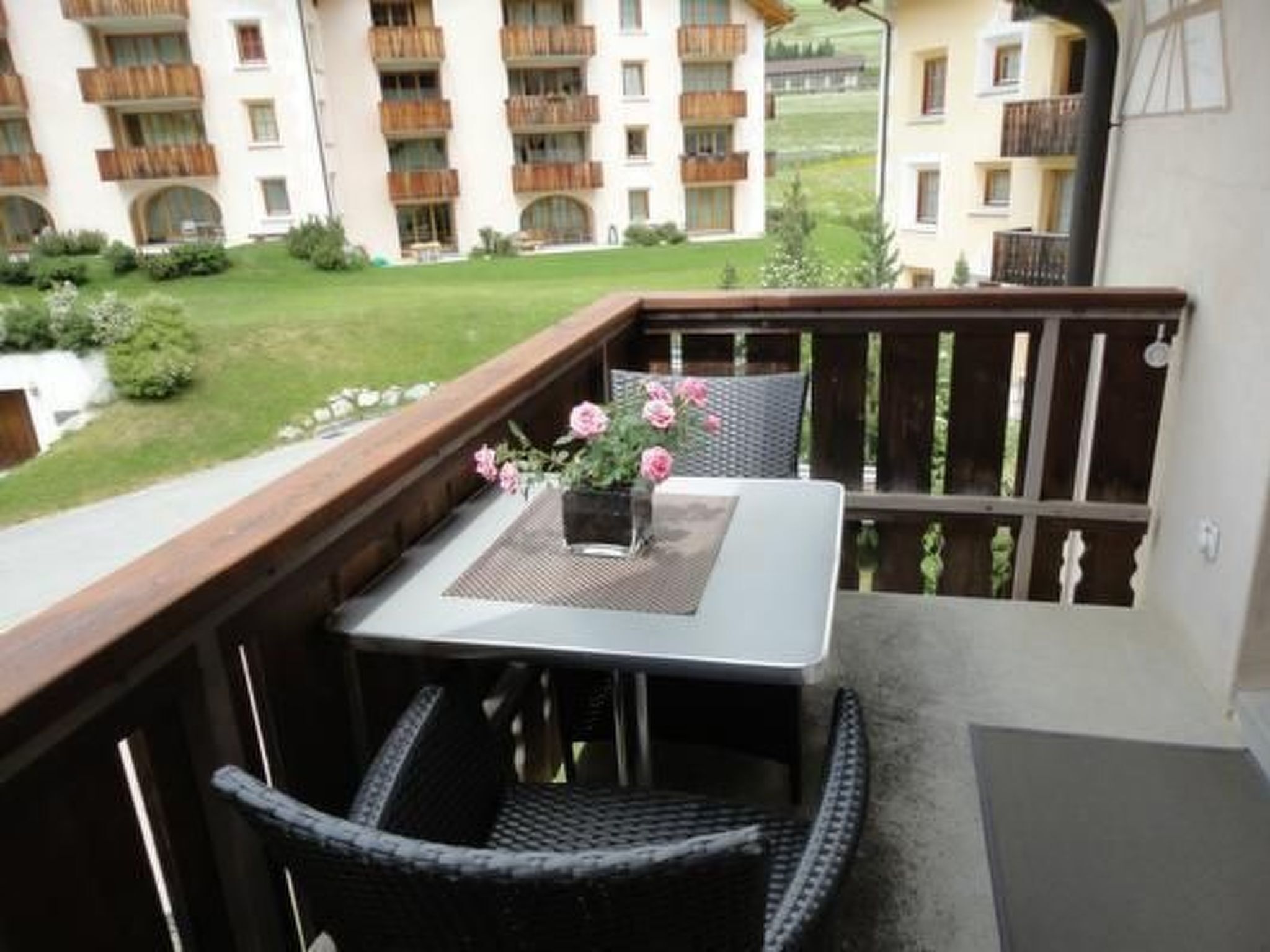 Un balcon avec une table et des chaises, un pot de fleurs orné de fleurs roses et une vue sur un immeuble doté de balcons.