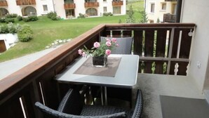 Uma varanda com mesa e cadeiras, um vaso com flores cor-de-rosa e vista para um edifício com varandas.