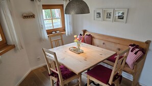 Un coin repas avec une table en bois, des chaises et un banc. Il y a une fenêtre avec des rideaux, un luminaire suspendu et des tableaux encadrés au mur.