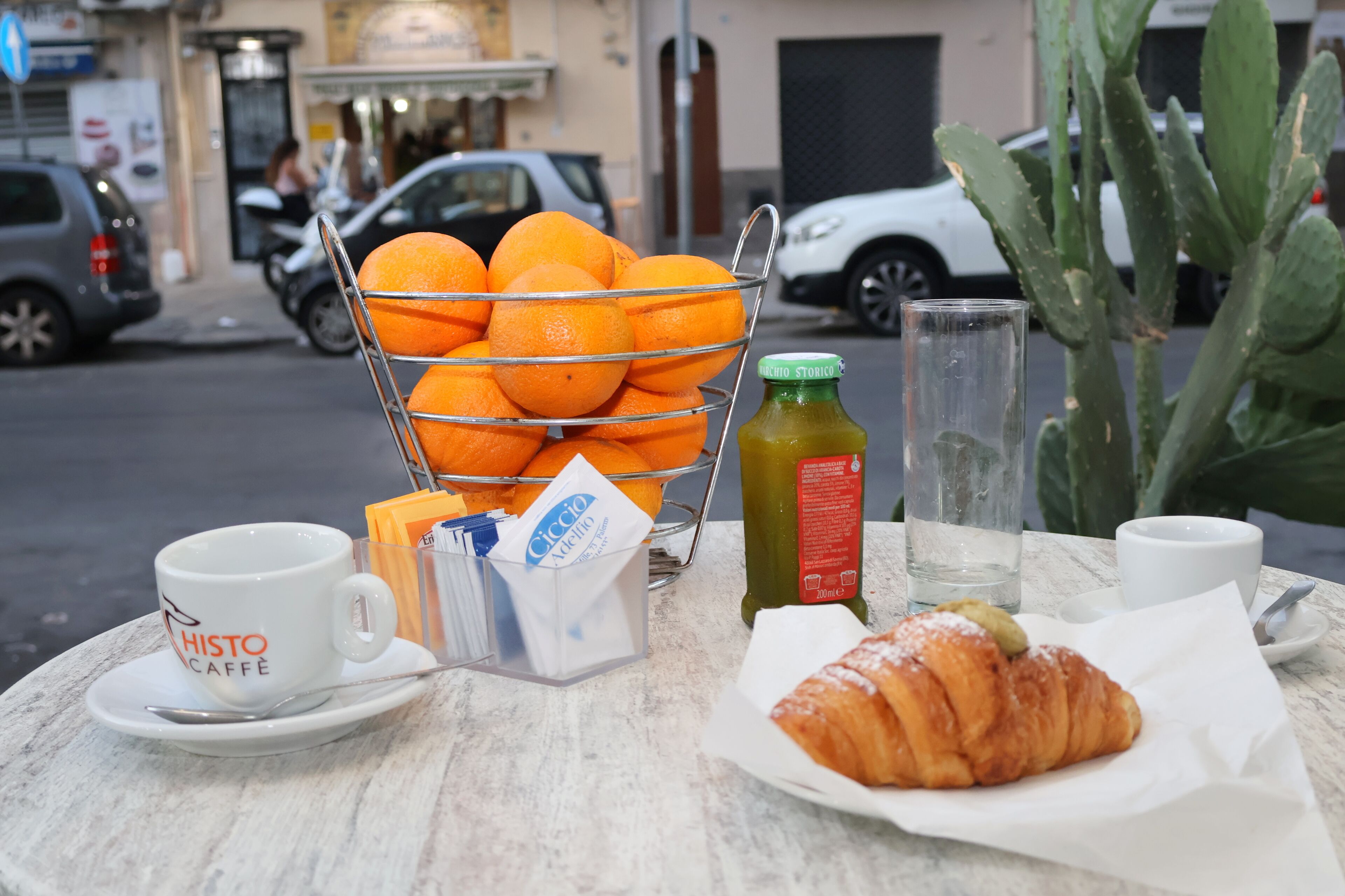 A table with a coffee cup, a basket of oranges, a bottle of juice, a glass of water, and a croissant.