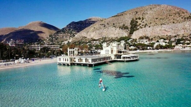 A large, white, multi-story building with a waterfront view, surrounded by clear blue water and a mountainous backdrop. - B&B Central Station  (Palermo)