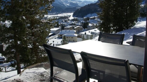 Uma mesa de jantar com cadeiras, montanhas cobertas de neve e uma cidade ao fundo.