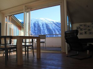 A room with a large window overlooking a snowy mountain. There is a wooden table and chairs in the foreground. - Sur Puoz 13 by Interhome (Samedan)
