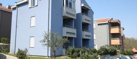 A multi-story residential building with balconies, a parking area, and a small garden.