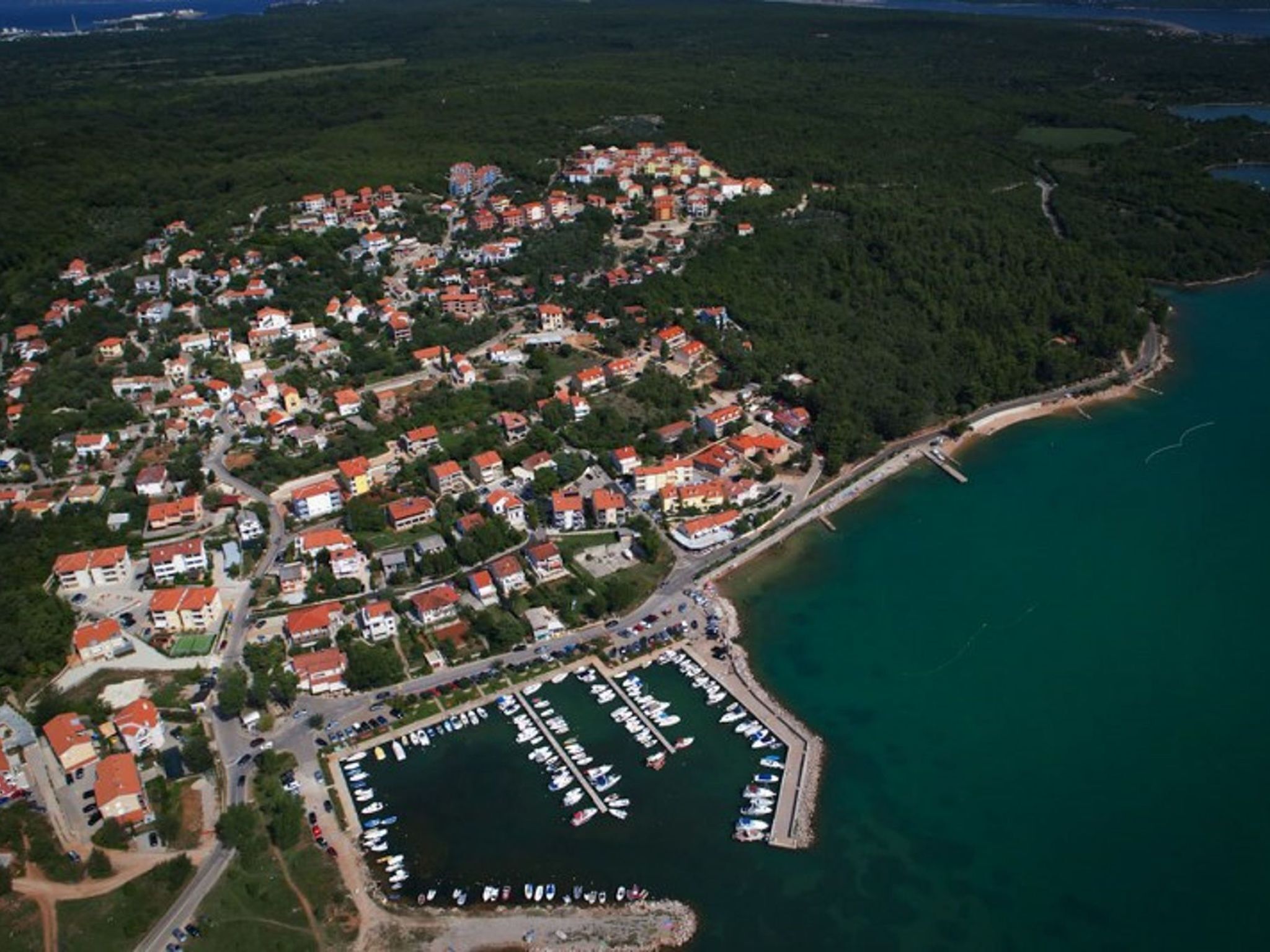 Vista aérea de un pueblo costero con edificios residenciales, un puerto deportivo y una carretera que lleva a la playa.