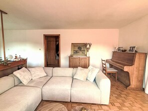 Een woonkamer met een hoekbank, een houten kast, een lijst met een landschap en een piano.