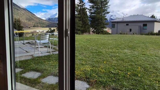 Une vue à travers une porte vitrée d’une cour verdoyante, avec une montagne en arrière-plan, un bâtiment et une terrasse avec des chaises.