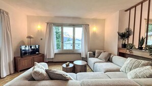 Een woonkamer met een hoekbank, een televisie op een houten kast en een raam met gordijnen.