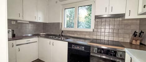 Une cuisine avec des meubles blancs, un lave-vaisselle, une cuisinière et une fenêtre donnant sur les arbres.