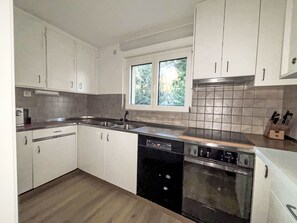 Een keuken met witte kasten, een vaatwasser, een fornuis en een raam met uitzicht op bomen.