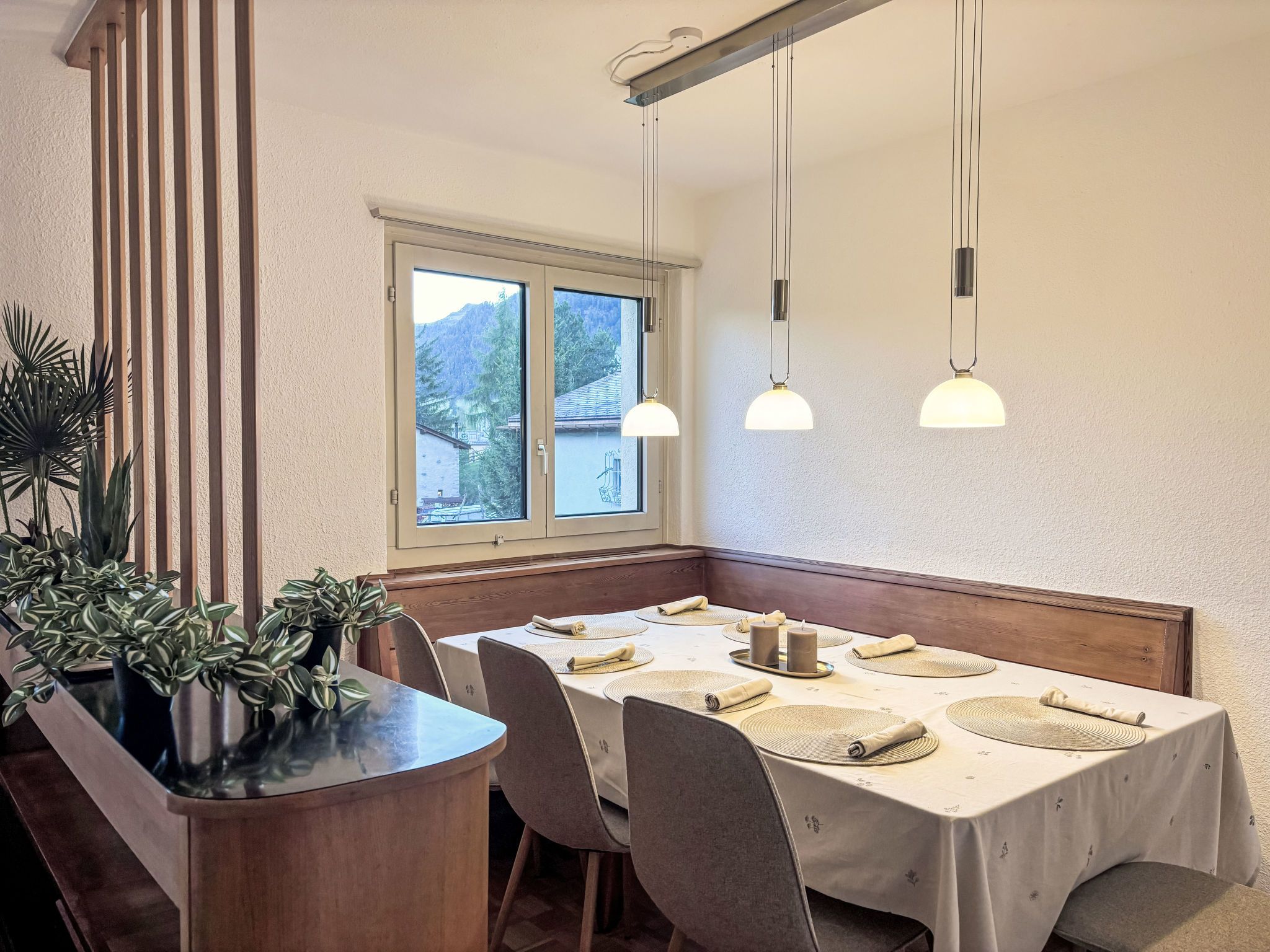 A dining area with a table set for four, a wooden bench, and a window with a view of a mountain.