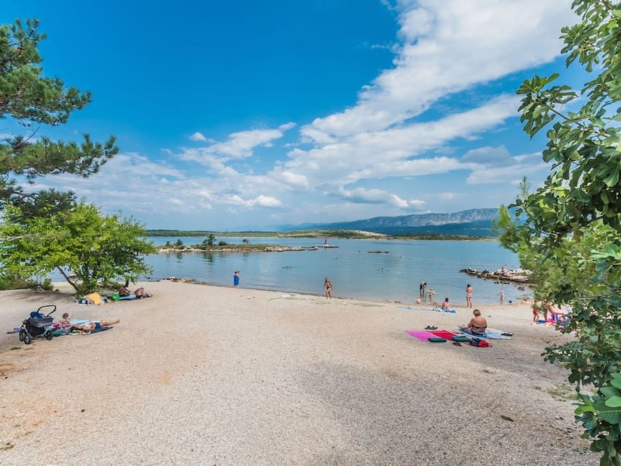 Une plage où des gens se prélassent au soleil et se baignent, entourée d’arbres et avec des montagnes en arrière-plan.