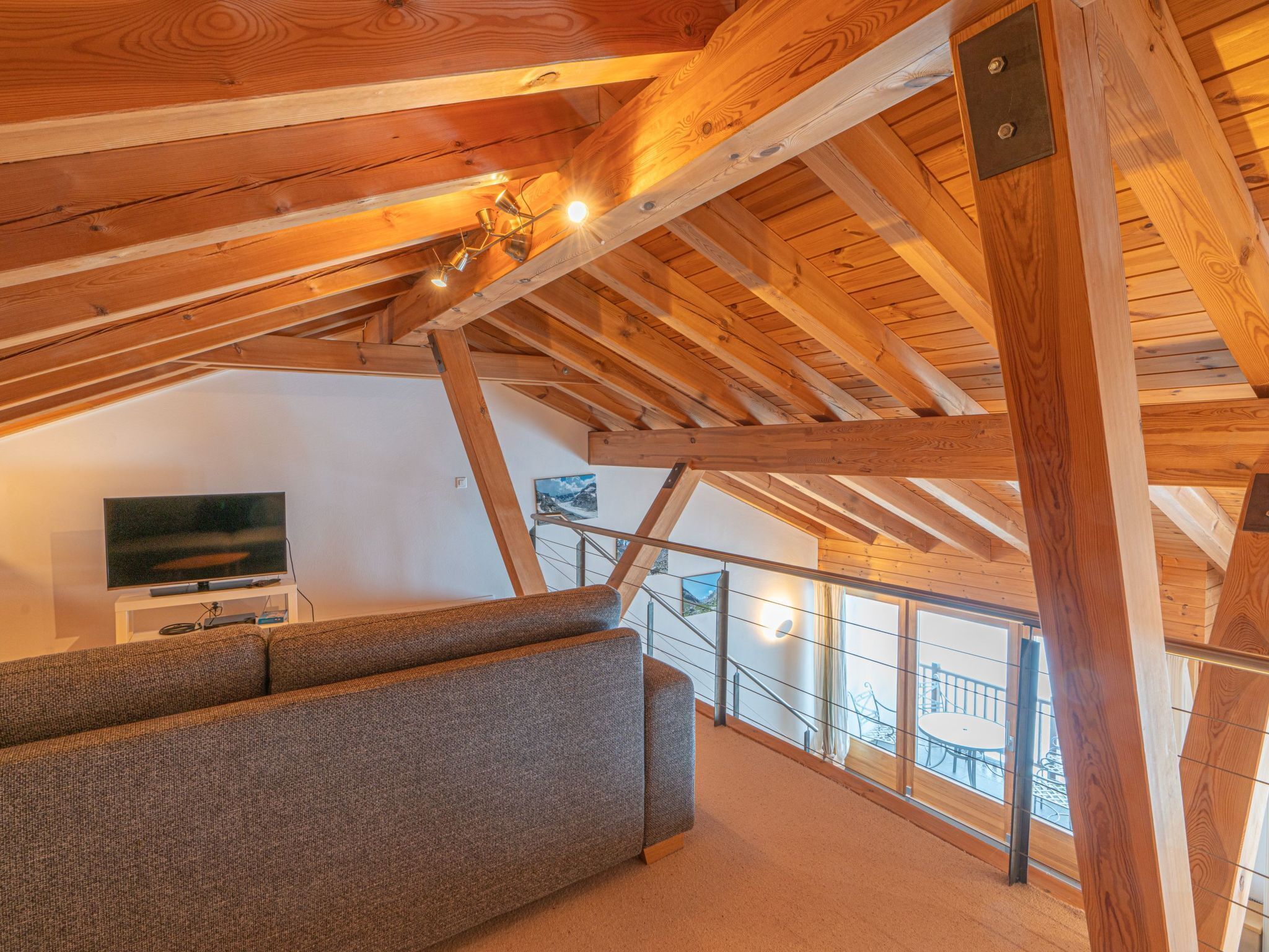 A loft-style room with exposed wooden beams, a sofa, a flat-screen TV, and a balcony with a railing.