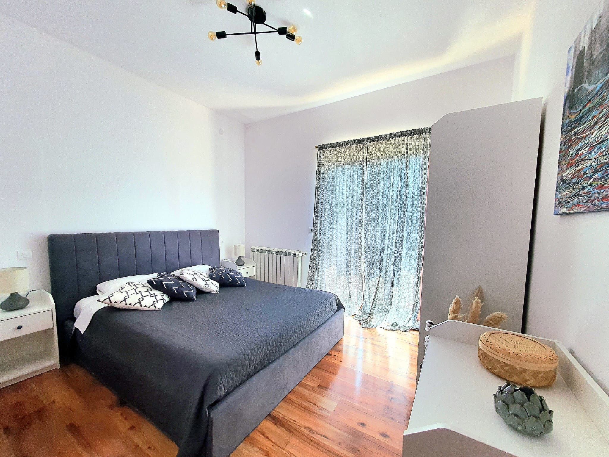 Una camera da letto moderna con un letto grande, un comodino, una lampada, un termosifone, una finestra con tende e un piccolo cesto decorativo.