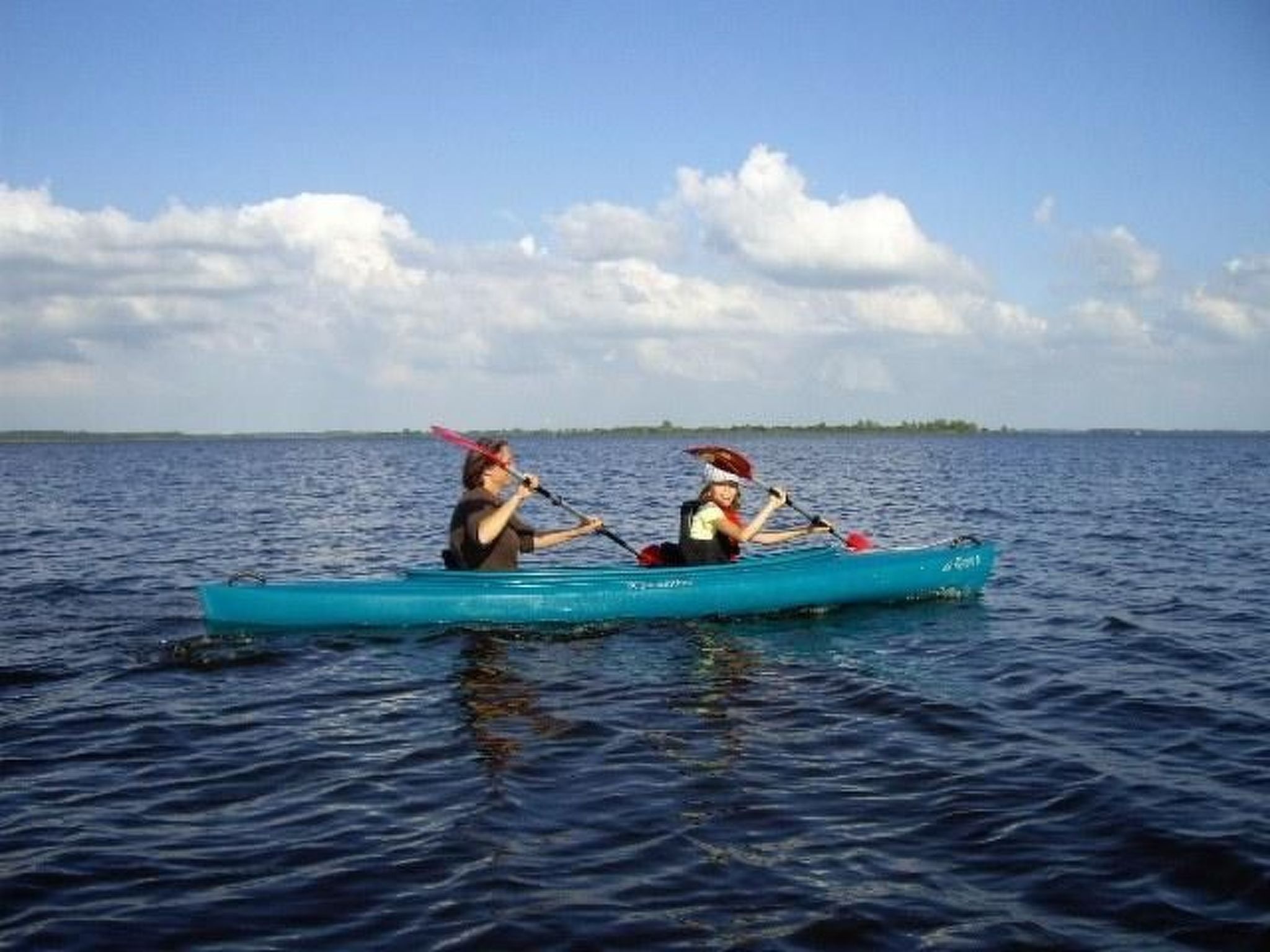 Deux personnes pratiquent le kayak sur une étendue d’eau calme, sous un ciel dégagé, et on aperçoit la terre ferme au loin.