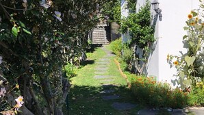 Ein schmaler Steinweg führt zu einem weißen Gebäude, umgeben von üppiger Vegetation und blühenden Pflanzen.