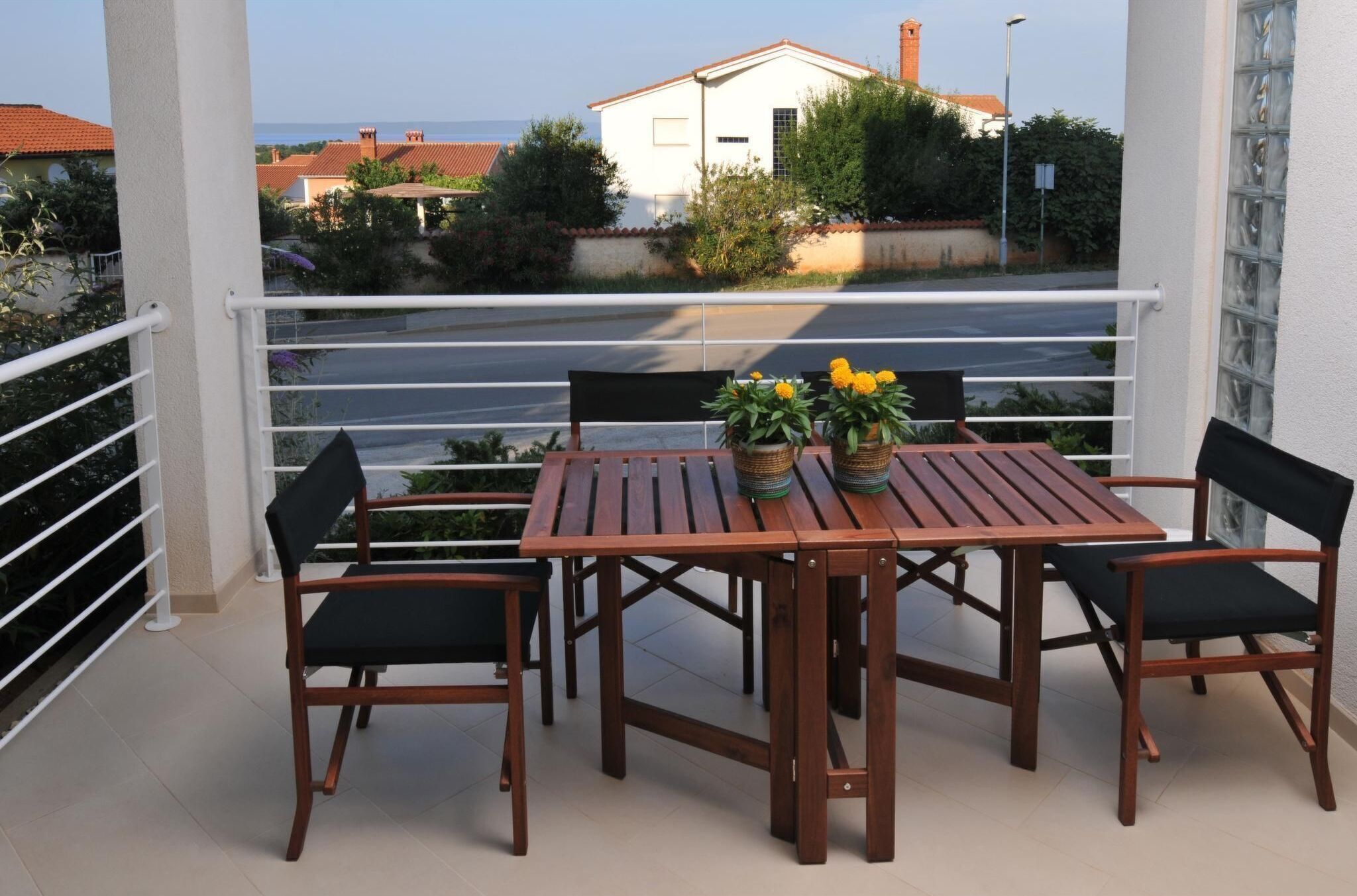 Una mesa de madera con cuatro sillas en un balcón, con vistas a casas y follaje al fondo.