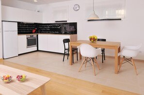 Una cocina moderna con armarios blancos, una encimera negra y una mesa de comedor de madera con cuatro sillas. Hay un reloj en la pared y un frutero sobre la mesa.