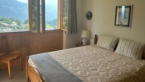 Un dormitorio con una cama grande, una silla de madera, una mesita de noche con lámpara, un cuadro enmarcado en la pared y una ventana con cortinas.