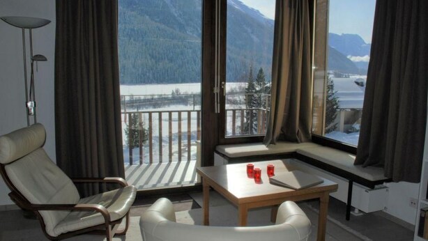 Una habitación con vistas a montañas y un lago, un sofá, una mesa con vasos rojos y una lámpara.