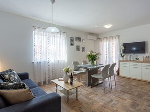 Ein modernes Wohnzimmer mit Sofa, Esstisch mit Stühlen, einem an der Wand montierten Fernseher und einer Küchenzeile inklusive Schrank und Fernseher.