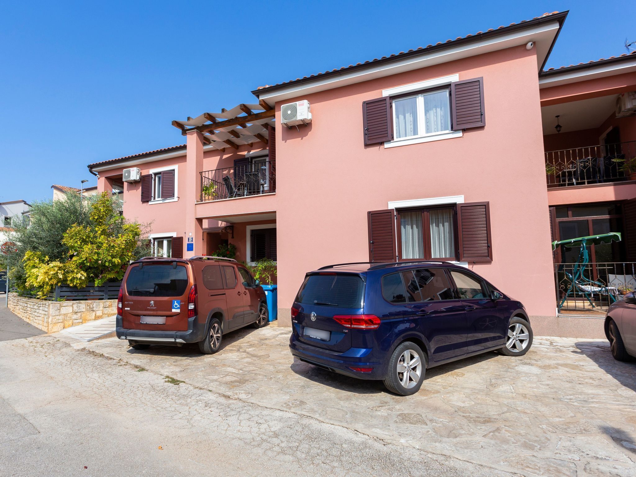 Un immeuble de deux étages avec une façade rose, un balcon doté d’une rambarde en métal et deux voitures garées.