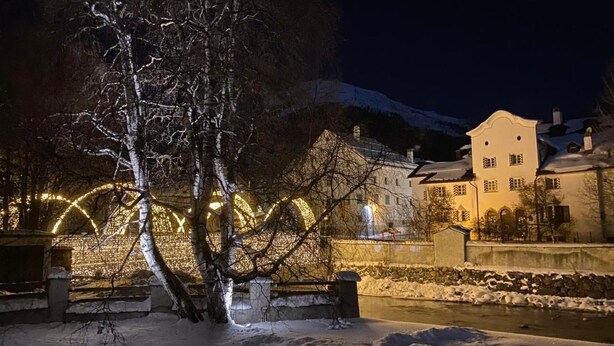 Zona nevada al aire libre con un árbol en primer plano, un muro de piedra y un edificio con arcos iluminados al fondo.