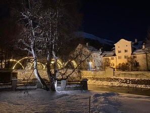 A snow-covered outdoor area with a tree in the foreground, a stone wall, and a building with illuminated arches in the background. - Chesa Engel by Interhome (La Punt-Chamues-ch)