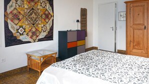 Um quarto com uma cama, um armário colorido, um enfeite na parede e uma cadeira de vime.