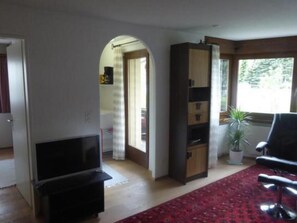 Un salon avec un téléviseur, une bibliothèque, une moquette rouge et une fenêtre donnant sur des arbres.
