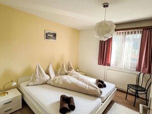 Une chambre à coucher comprenant un lit, des oreillers, une table de chevet, une chaise, une fenêtre avec des rideaux et un luminaire suspendu.