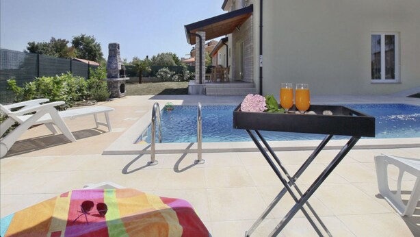 Uma área de piscina com espreguiçadeiras, uma mesa com bebidas e um guarda-sol colorido.