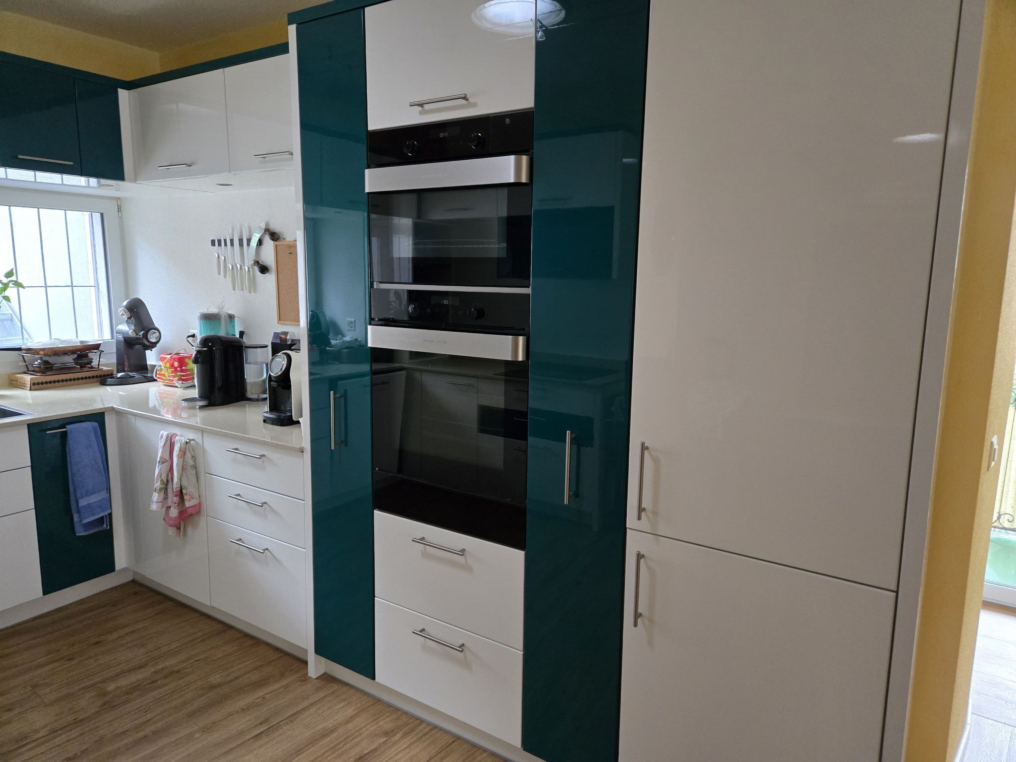 A modern kitchen with white and teal cabinets, a built-in oven, and a coffee maker on the counter.