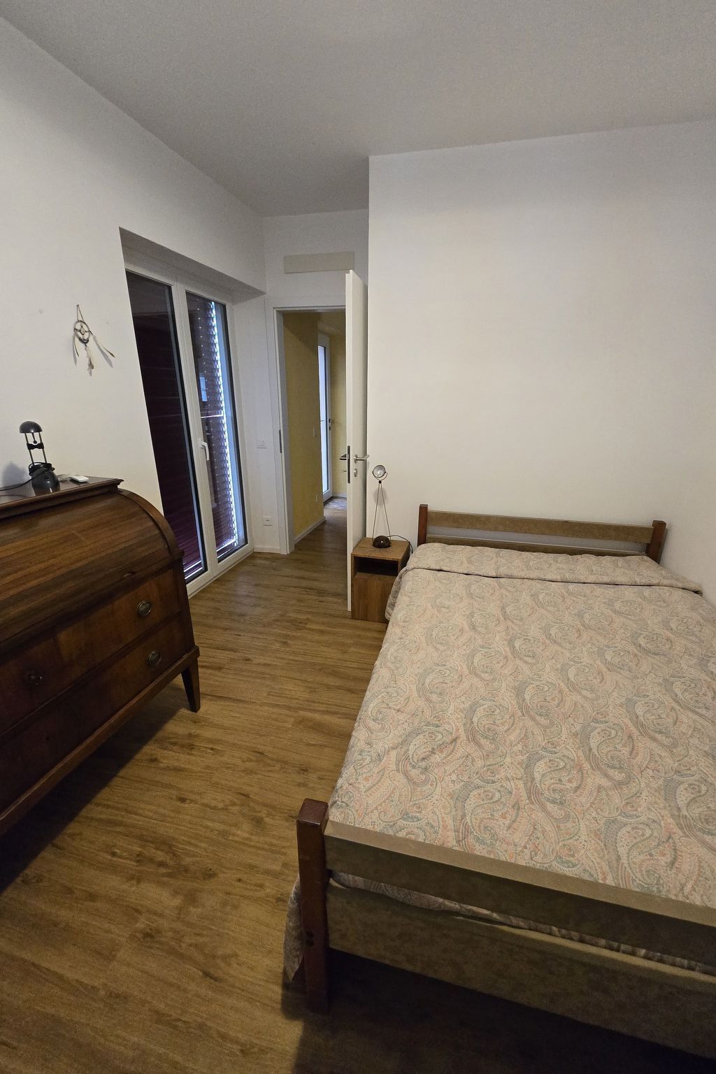 Un dormitorio con cama de madera, cómoda y mesita de noche. Se ve una puerta que conduce a otra habitación y una ventana con persianas.