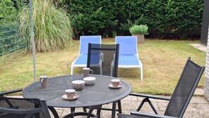 Une terrasse extérieure avec une table ronde et quatre chaises, un parasol bleu et une haie en arrière-plan.