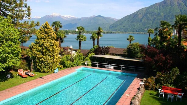 En swimmingpool med kristallklart blått vatten, omgiven av frodig grönska och med utsikt över en sjö där berg reser sig i bakgrunden.