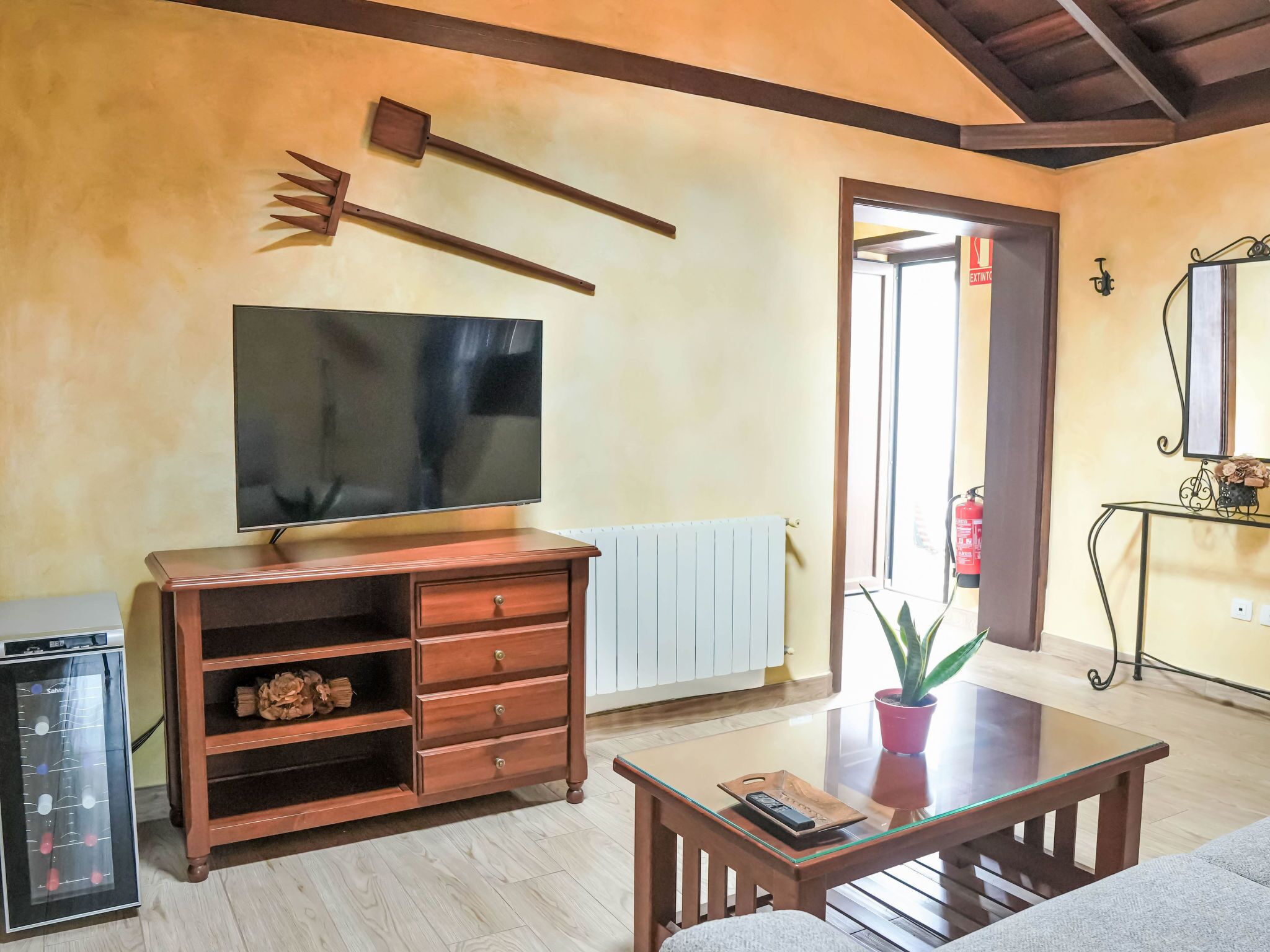 A living room with a TV, wooden furniture, a glass coffee table, and a potted plant.