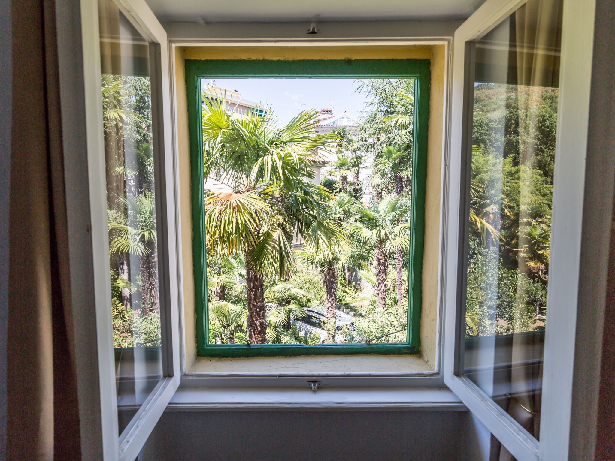 A window with a green frame, offering a view of a lush garden with various trees and plants.