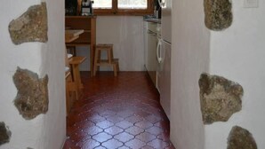 A narrow hallway with a tiled floor leading to a kitchen area with a window, wooden furniture, and stone wall accents. - Bos-cha by Interhome (La Punt-Chamues-ch)