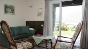 Een woonkamer met een groene bank, twee houten stoelen, een bijzettafeltje met een boek en een vloerkleed met patroon.