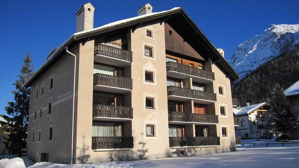 Un edificio de varios pisos con balcones, una pista de esquí al fondo y nieve en el suelo.