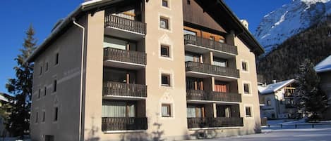 A multi-story building with balconies, a ski slope in the background, and snow on the ground.