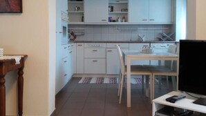 Eine kompakte Küche mit weißen Schränken, einem Esstisch mit Stühlen, einem Fernseher und einem kleinen Tisch mit einer Lampe.
