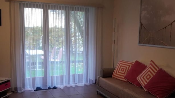 Ein Wohnzimmer mit einem beigen Sofa, gemusterten Kissen, einem großen Fenster mit durchscheinenden Vorhängen und einem Fliesenboden.