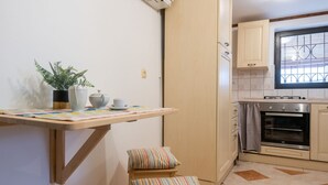 Una cocina compacta con horno integrado, una ventana con reja de metal y un pequeño comedor con mesa y banco.